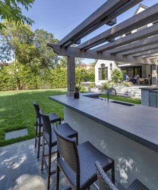 Shaded garden bar ideas under a gray pergola with dark gray wicker bar stools in a lawned garden space.