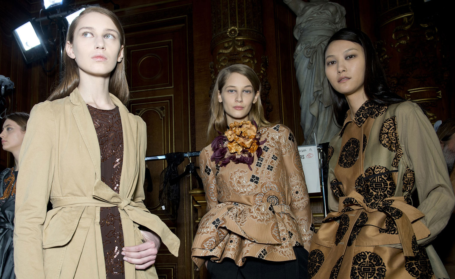One woman in a cream coat over a purple dress, and two women wearing tan coats with baroque detailing
