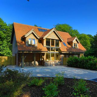 view of the front exterior of a self build large 3 storey house with oak frame and orange tones, a large stone paved driveway bordered by garens