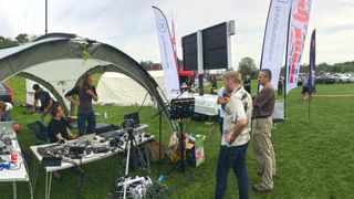 Live Commentators with microphones and show runners in tents at the outdoor International Drone Day in a field with flags in the background