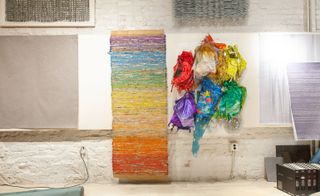 White brick wall, colourful finished tapestry hung in wall, bunch of recycled balloons in a variety of colours, neutral stone floor, wired plug in wall socket, samples and brown files to the right corner of the shot