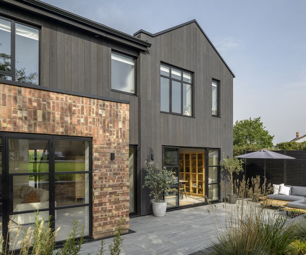 The rear exterior of a house extension and renovation project. There is a large paved patio which the kitchen opens out to .The cladding is contemporary black timber and brick