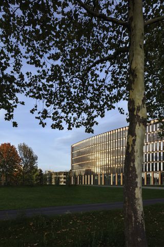 The Freiburg Town Hall by Ingenhoven Architects reads like an eco-coliseum. A large many leveled building with silver vertical panels between many vertical windows in front of a park.