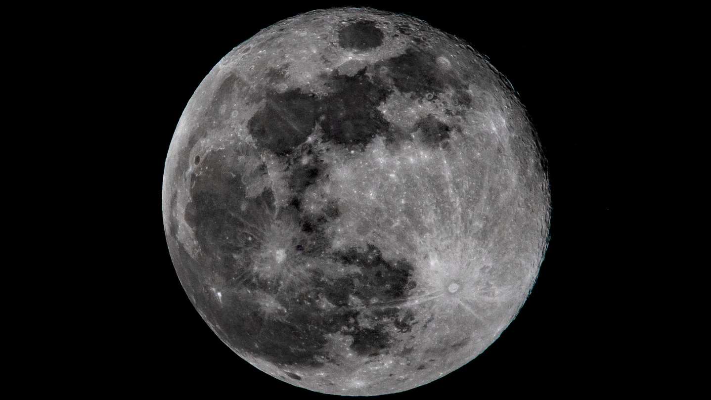 A full moon is shown against a black sky, with dark basaltic plains and bright lunar craters marking its ancient surface.