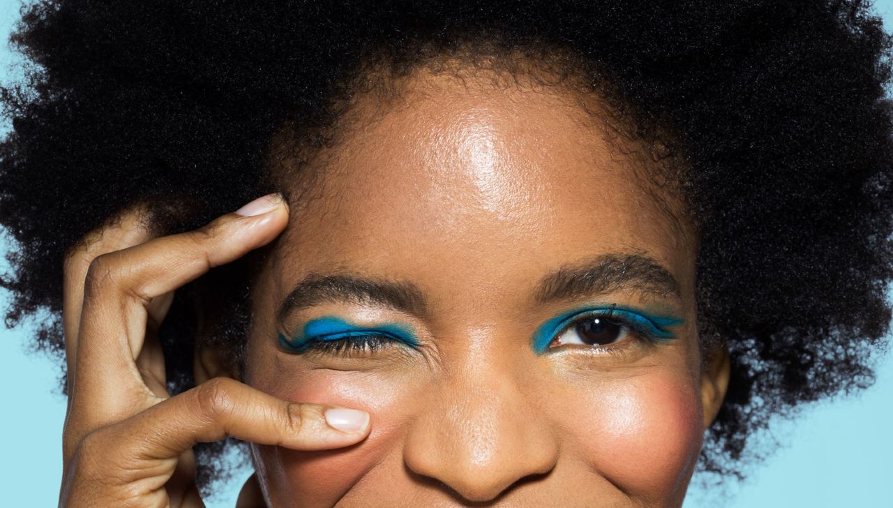 Close-up of young mixed-race woman looking to camera, smiling, wearing blue eye make-up, with hand near eye, with turquoise background