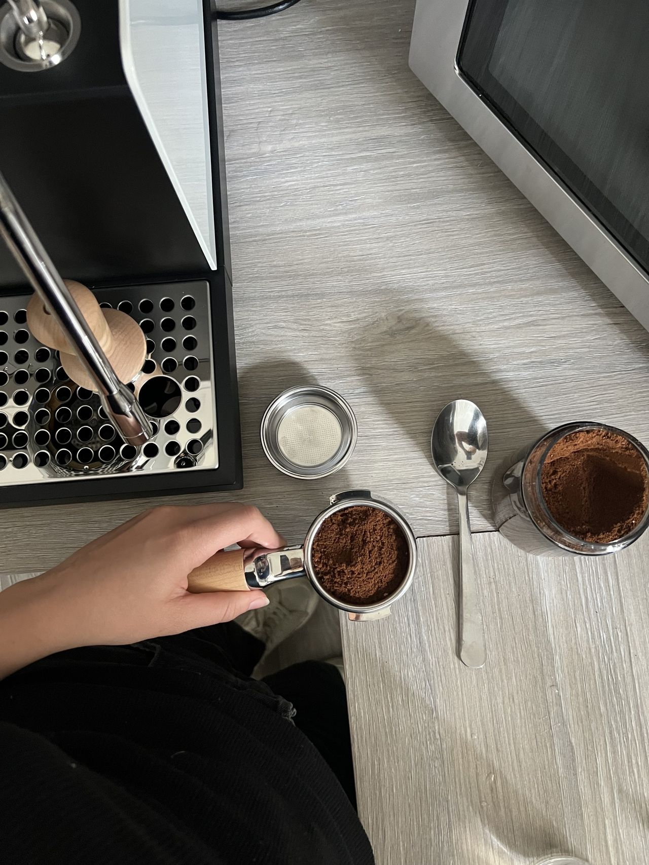 A hand holding a portafilter full of coffee grinds, and there is a container of coffee grinds next to it, as well as a fellow series 1 espresso machine.