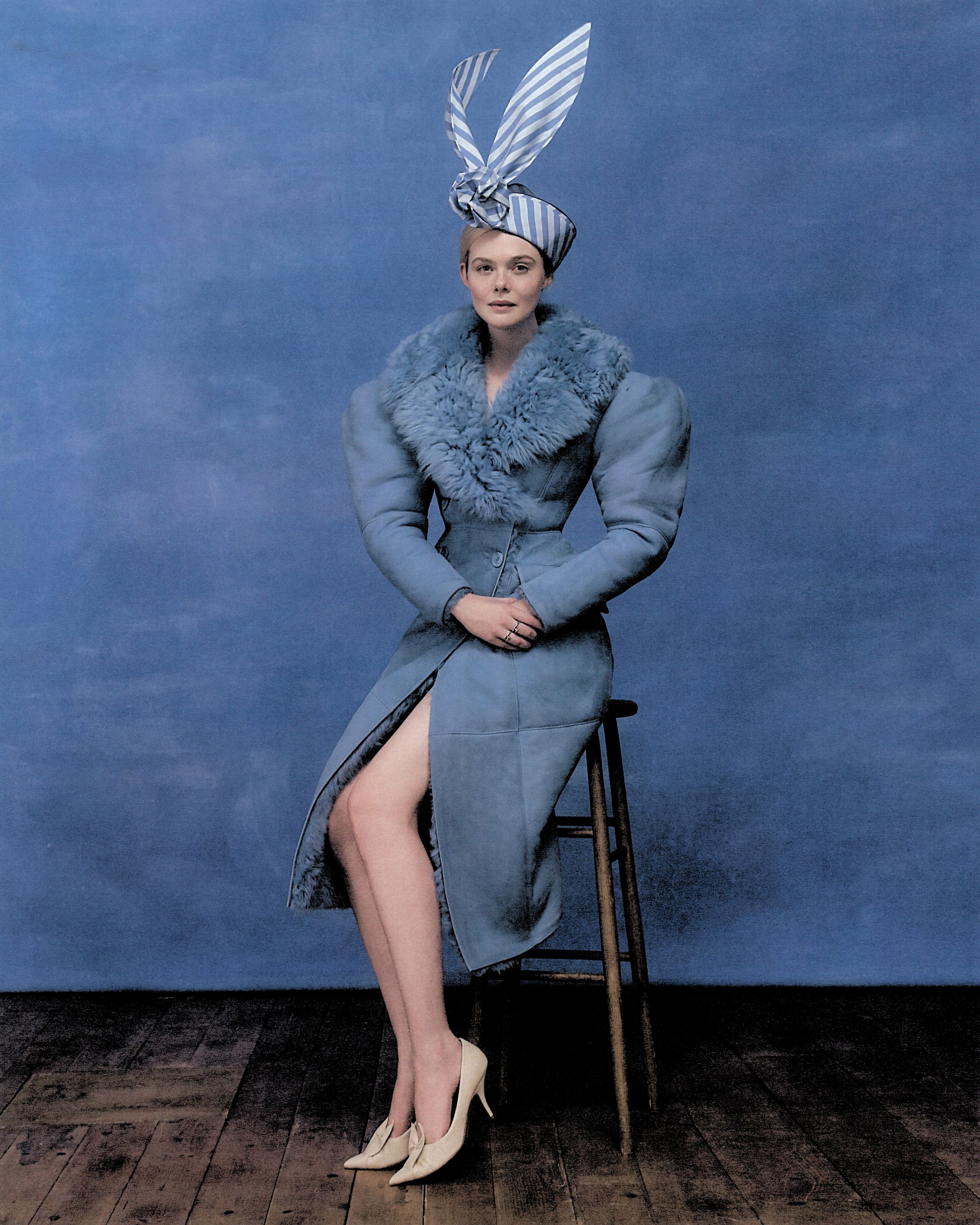 Elle Fanning shot by Szilveszter Makó. She is sitting on a wooden stool in front of a painted blue backdrop. She is wearing a blue Gucci coat with faux fur trim collar, a blue and white stripe hat, and cream pointed-toe heels.