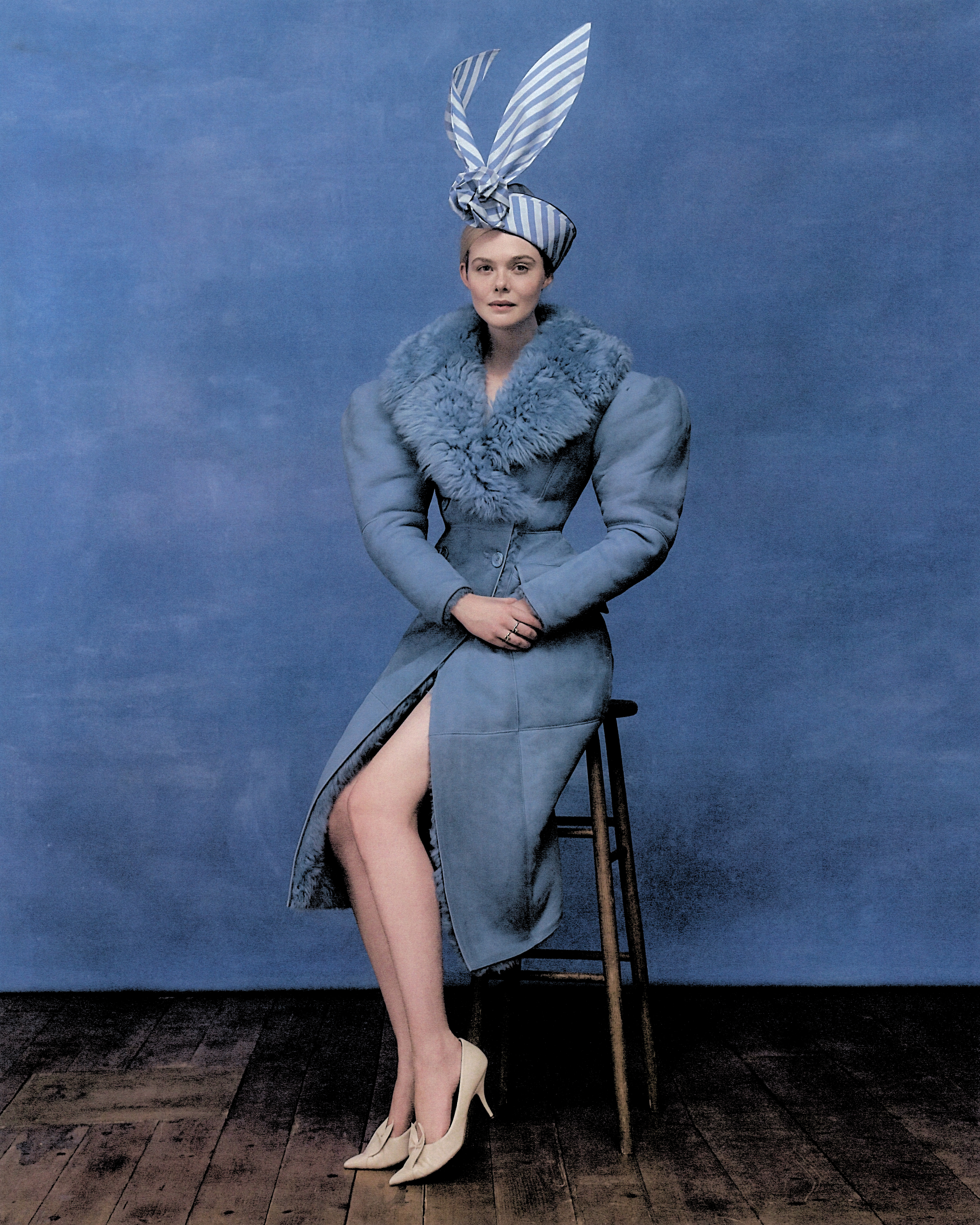 Elle Fanning shot by Szilveszter Mak&amp;oacute;. She is sitting on a wooden stool in front of a painted blue backdrop. She is wearing a blue Gucci coat with faux fur trim collar, a blue and white stripe hat, and cream pointed-toe heels.