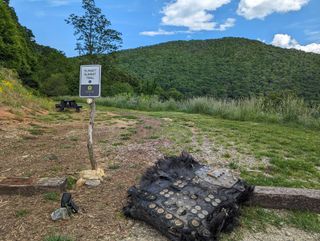 a large black piece of fiberglass covered in metal bolts and plates lies on the ground beside a trail leading into a forest. mountains can be seen rolling in the distance