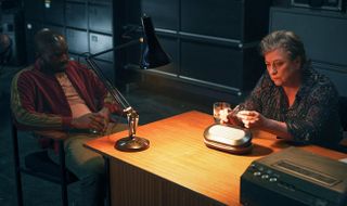 George (Paapa Essiedu) and Wes (Caroline Quentin) sit in a darkened office, the only light coming from a lamp on the desk in front of Wes. George is sitting on the opposite side, slumped back in a chair, while Wes is staring intently at a pen-like device in her hands