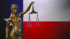 Close-up of a small bronze statuette of Lady Justice before a flag of Texas.