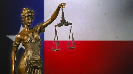 Close-up of a small bronze statuette of Lady Justice before a flag of Texas.