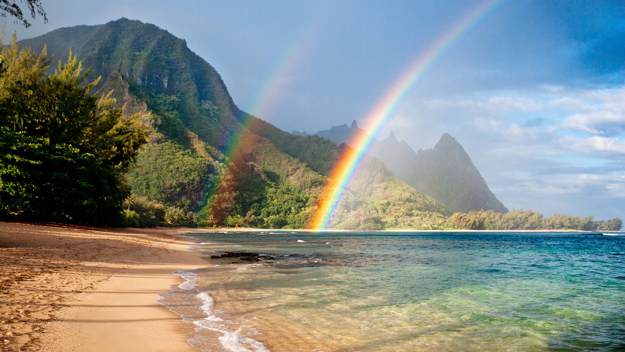 A double rainbow is seen over a beach with a mountainside in the background