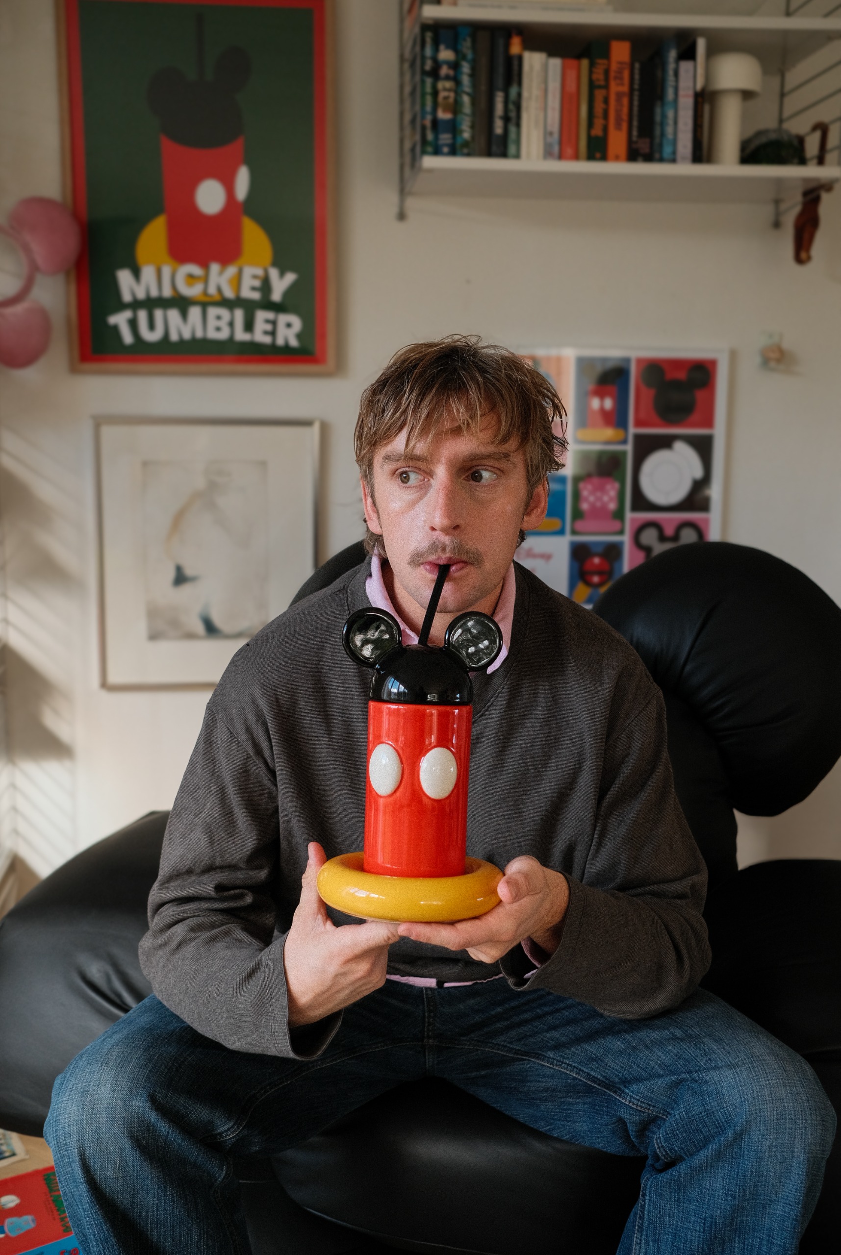 A young man with blonde hair, a gray jumper over a pink shirt, and blue denim jeans drinks from a Mickey Mouse-inspired tumbler sitting in a colorful room on a black leather sofa.