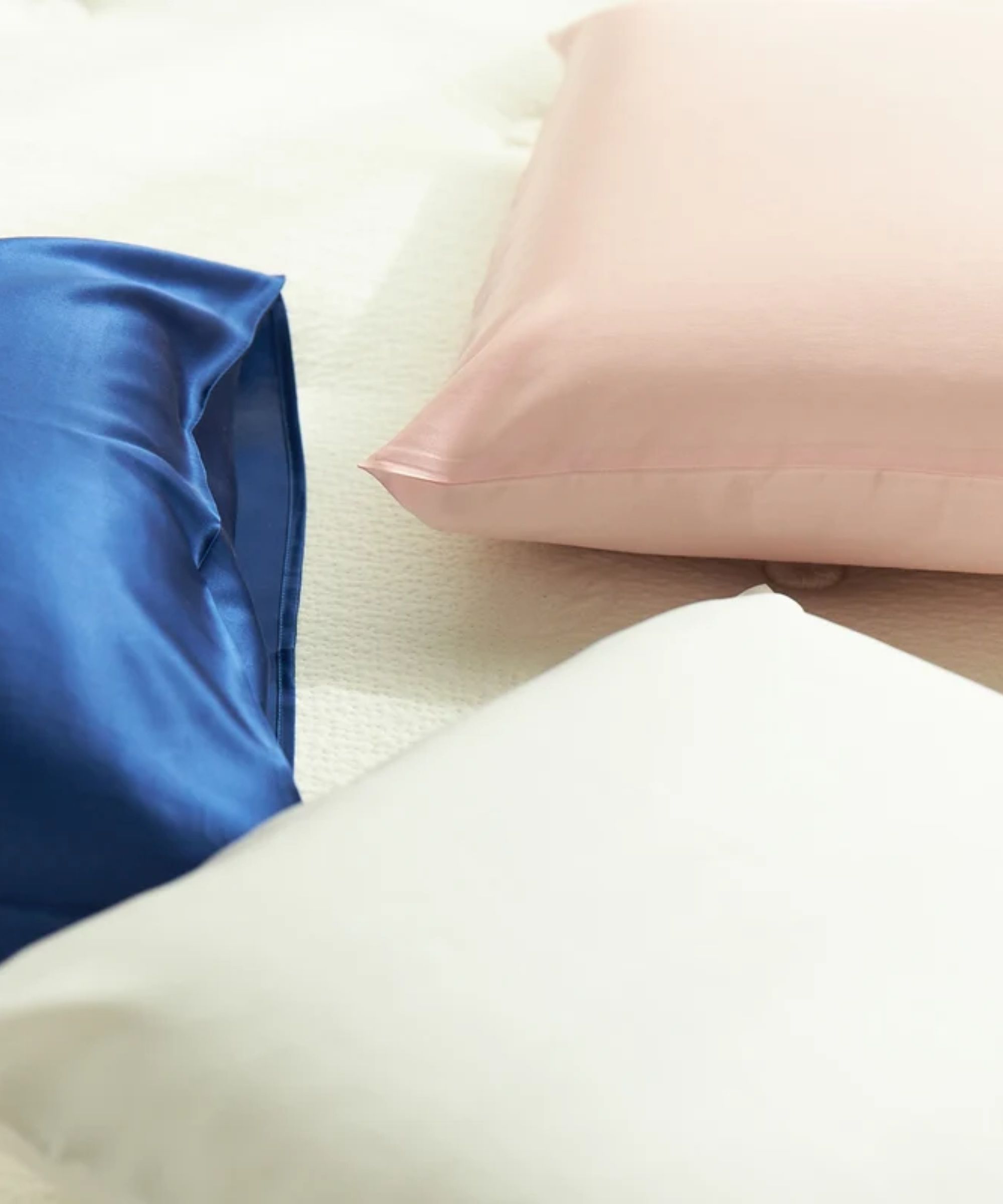 Three Avocado silk pillowcases pictured in different colors on pillows, on top of a soft white surface