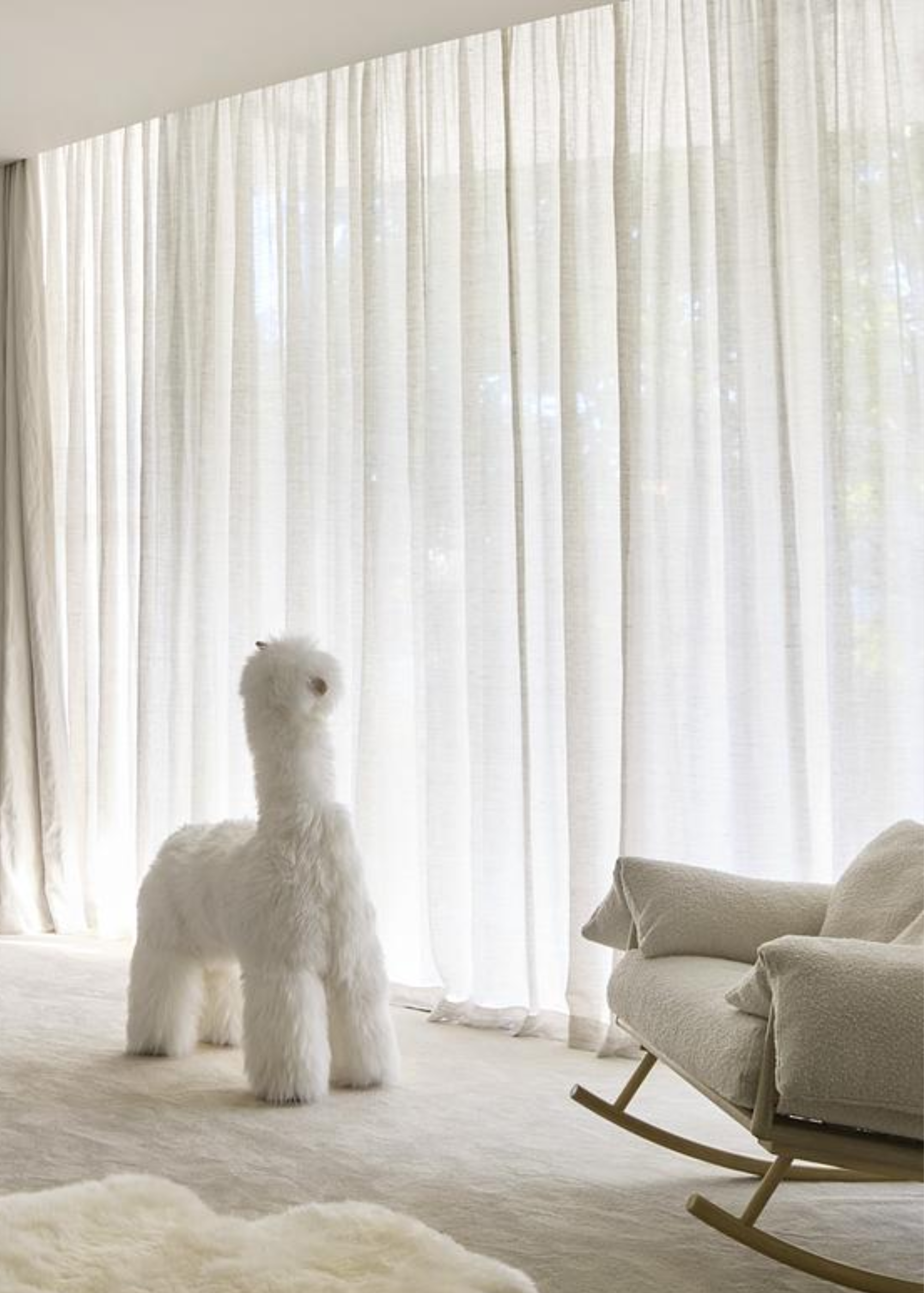 A nursery with bright white tones and a toy placed beside a rocking chair. There are also sheer linen curtains hanging in the room with natural light flowing in.