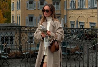 Woman wearing a long tan coat with white turtleneck sweater.