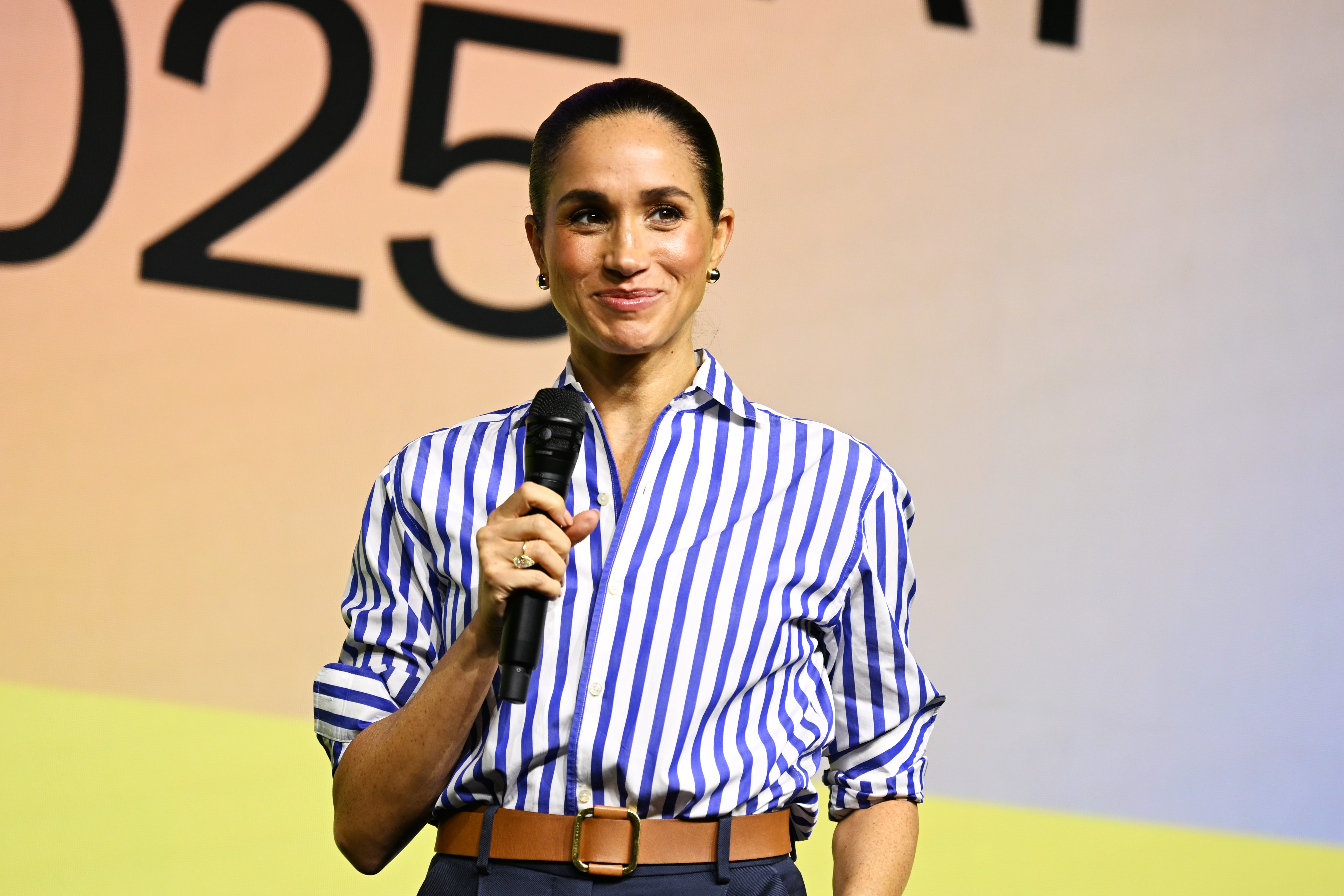 Meghan Markle wearing a blue striped shirt and blue pants speaking into a mic