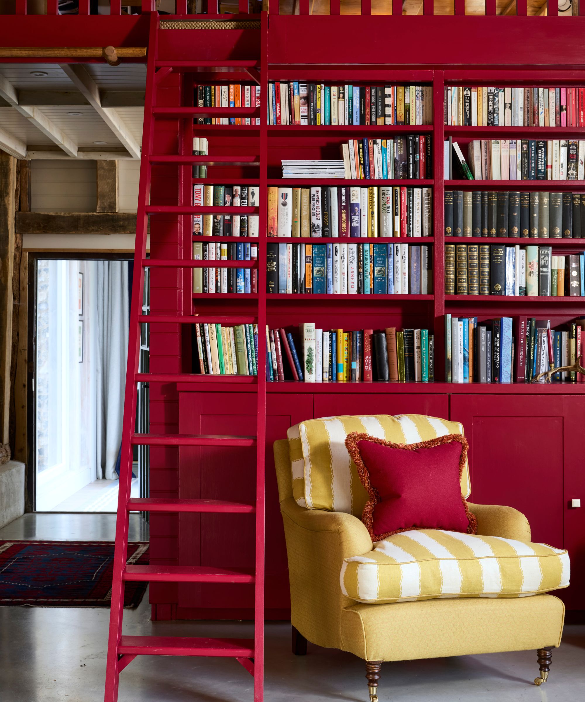 a rustic beamed barn conversation with bespoke red painted bookcase joinery, a loft ladder, and a striped yellow armchair