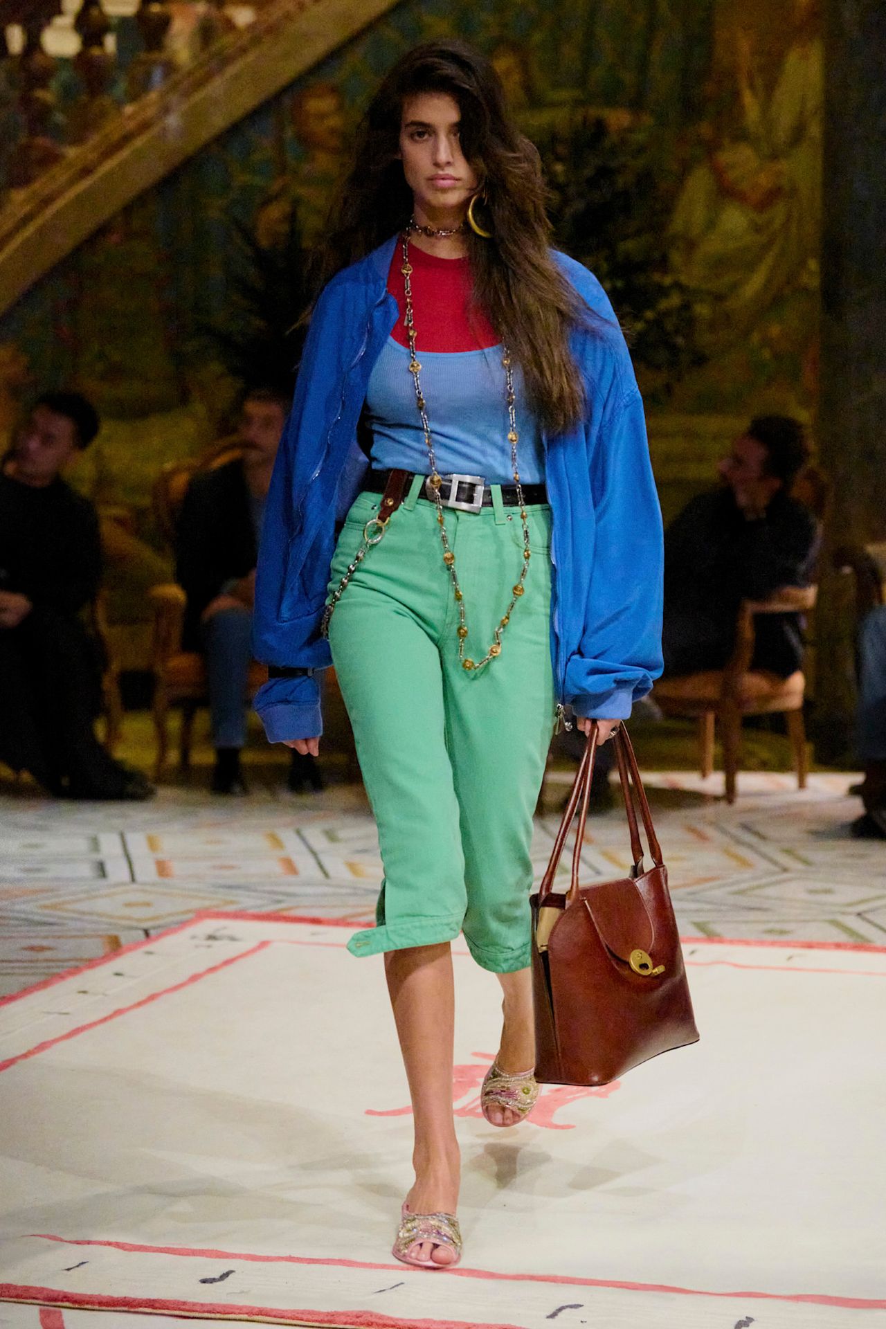 Layering staples for spring 2026 is shown in a photo from Versace's runway show, with a model walking while wearing a bright blue button-down over a red t-shirt, both layered under a blue tank top. The layers are paired with green pedal pushers, a black belt, tan sandals, a dark brown bag, and a long necklace.