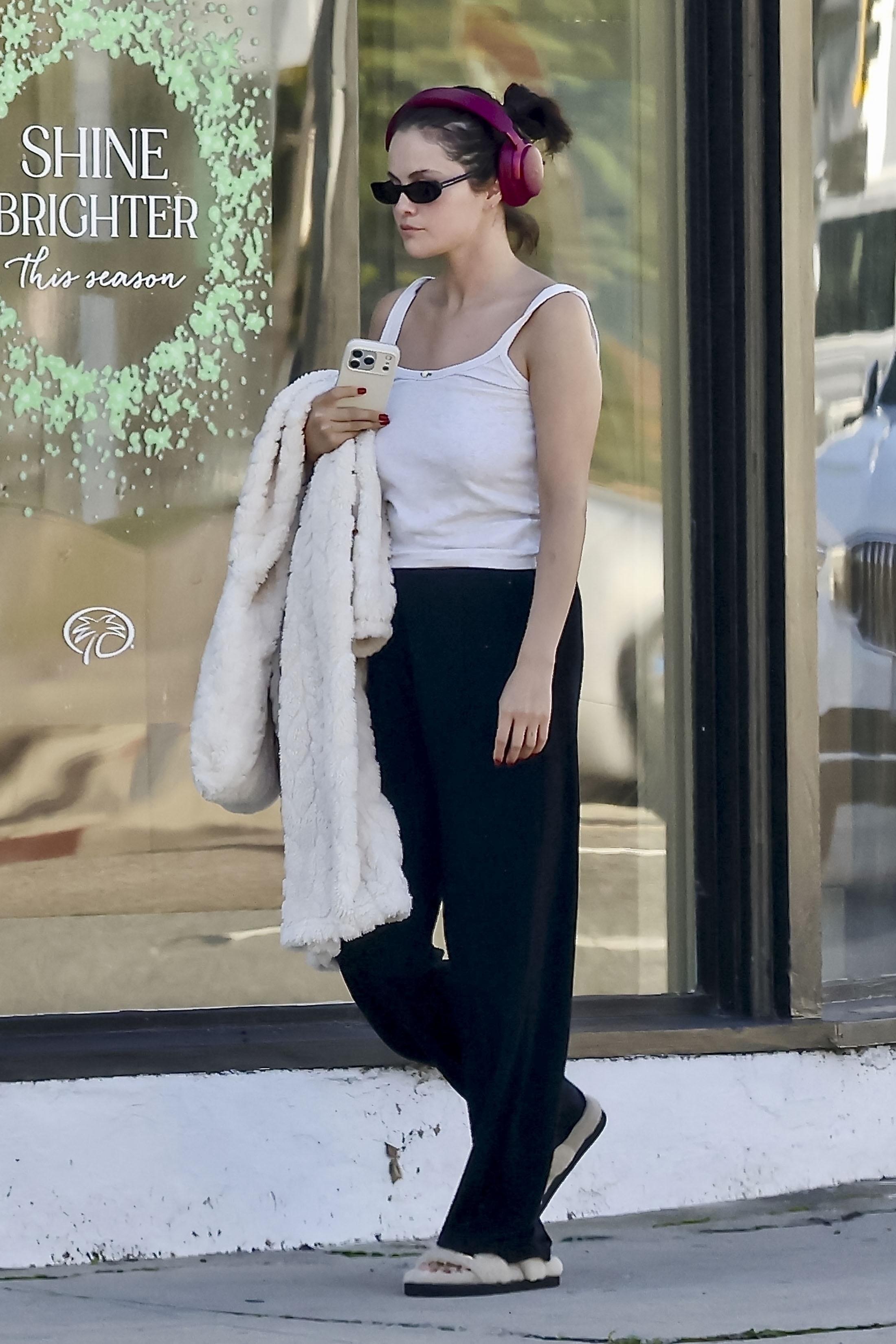 Selena Gomez was seen stepping out in Beverly Hills after a visit to Palm Beach Tan, sporting a relaxed yet stylish look. The singer and actress wore a white tank top, black sweatpants, and beige fuzzy slippers, accessorized with a cream-colored faux fur coat and pink headphones.