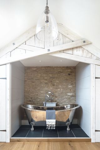 shiny steel bath tub in alcove with stone wall behind and white wood panels around and black floor tiles