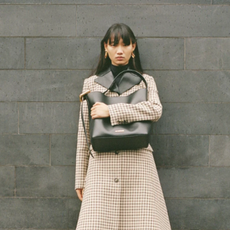 Woman in checked coat and gold earrings holding Selfridges bag with grey brick background
