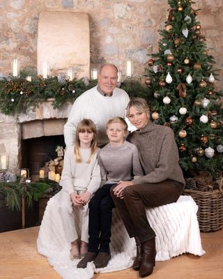 Prince Albert, Princess Charlene, Prince Jacques, Princess Gabriella sitting in front of a fireplace and Christmas tree on a white blanket