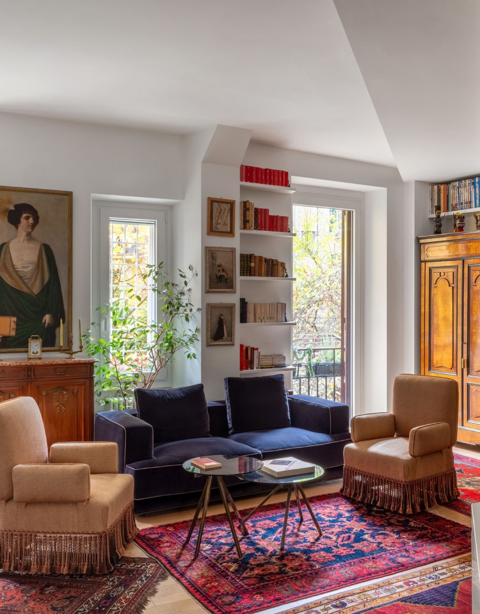 Image of an eclectic living room with two fringe-trimmed accent chairs, a navy sofa, multiple red rugs, and lots of art on the walls.