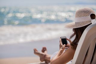 A woman sits on a chair at the beach, wearing a wide-brimmed hat, checking her phone.