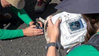 a researcher holds a white camera, which is focusing on a pair of hands shaping a pile of rocks and dirt on the ground