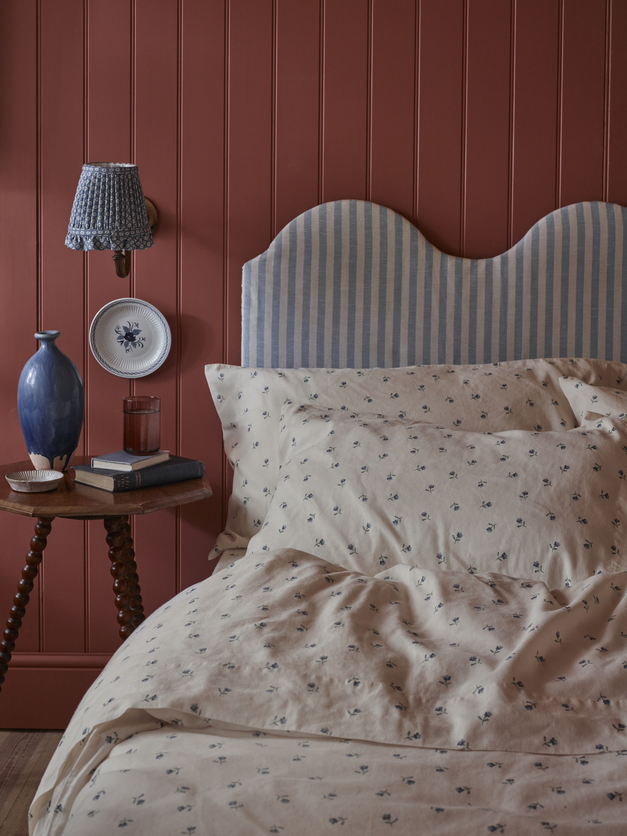 A bed that has a light blue and white striped, wavy headboard with blue and white, dainty floral bedding. The walls are red, and there is a small wooden side table beside the bed.