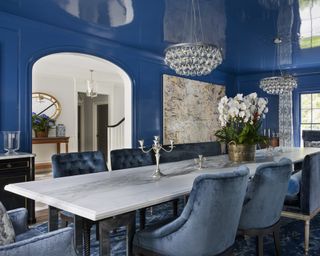 A dining room wall idea with blue gloss painted walls and ceiling, blue velvet dining chairs, artwork on one wall and a crystal chandelier on the ceiling