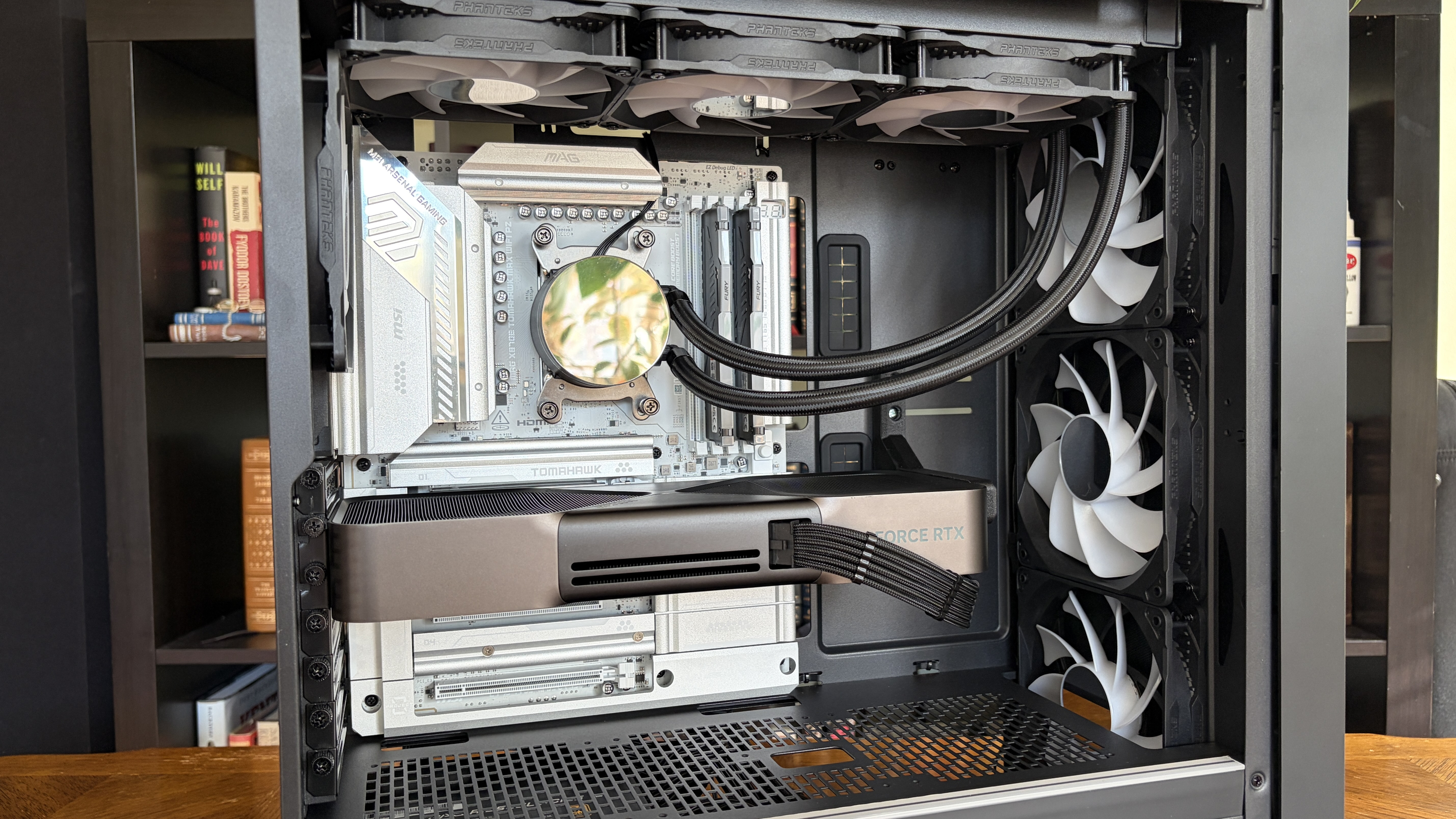A high-end Maingear MG-1 MK.II gaming PC open case with visible graphics card, liquid cooling tubes, and multiple cooling fans. Bookshelf blurred in the background.