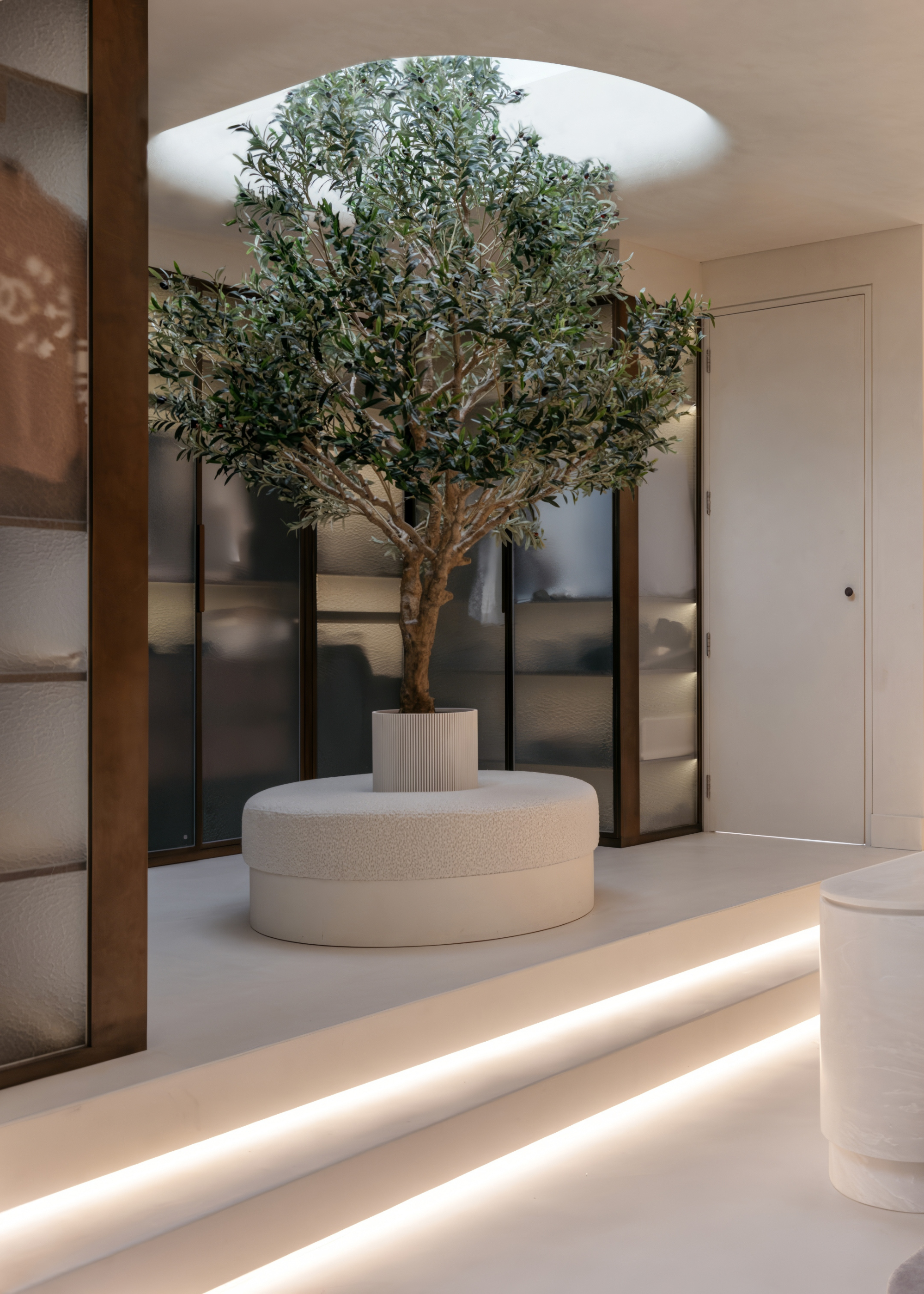 A minimalist bedroom with a large ribbed glass wardrobe by a round banquette seat with an olive tree in the middle