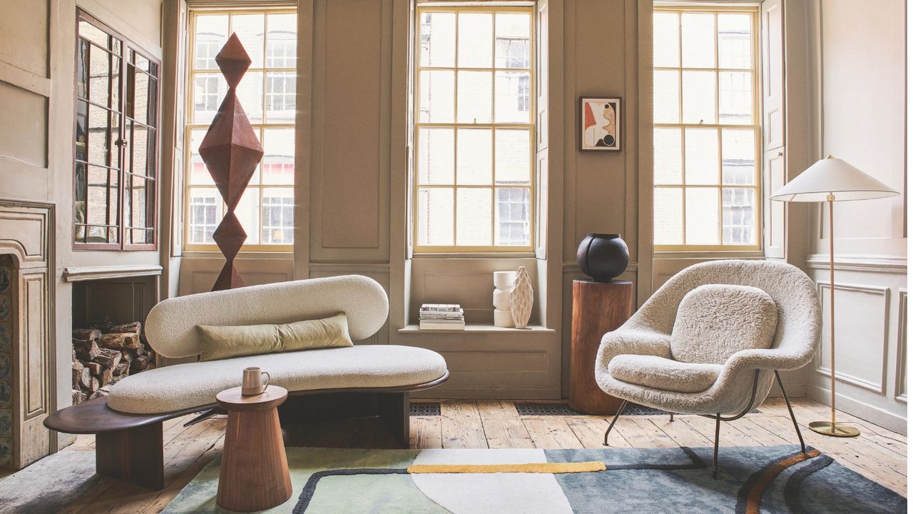 Bright apartment living room with curved modern boucle seating, window seats and three tall crittal windows facing an apartment building. There is a log fire and geometric rug