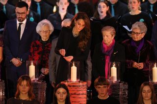 Kate Middleton wearing all black and lighting a candle in front of a crowd of people standing up