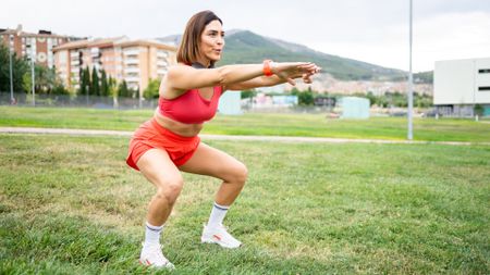 Woman in her 40s or 50s in red workout gear outdoors performing a squat on grass