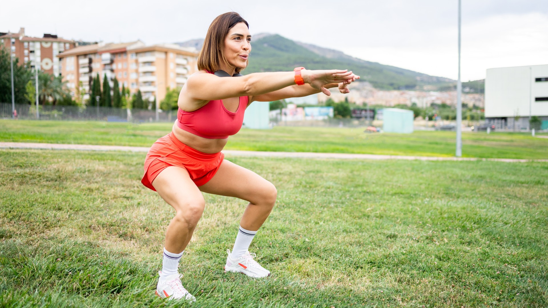 Woman in her 40s or 50s in red workout gear outdoors performing a squat on grass
