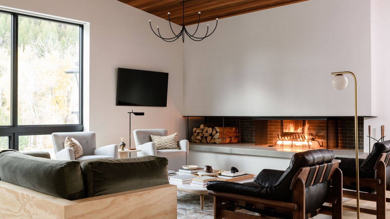 modern cabin interior with wood-burning fireplace, modern chandelier, leather accent chairs, and views of trees outside 