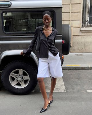 Paris-based fashion influencer Sylvie Mus poses in front of an SUV wearing a gold collar necklace, silk black button-down shirt, white Bermuda shorts, and black slingback heels