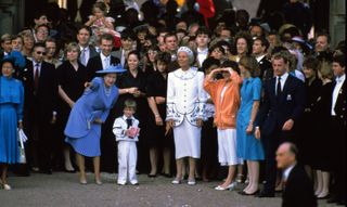 Queen Elizabeth wearing a blue skirt suit and pointing while talking to Prince William in a white sailor suit as they stand in front of a big group of guests standing outside Sarah Ferguson and Prince Andrew's wedding