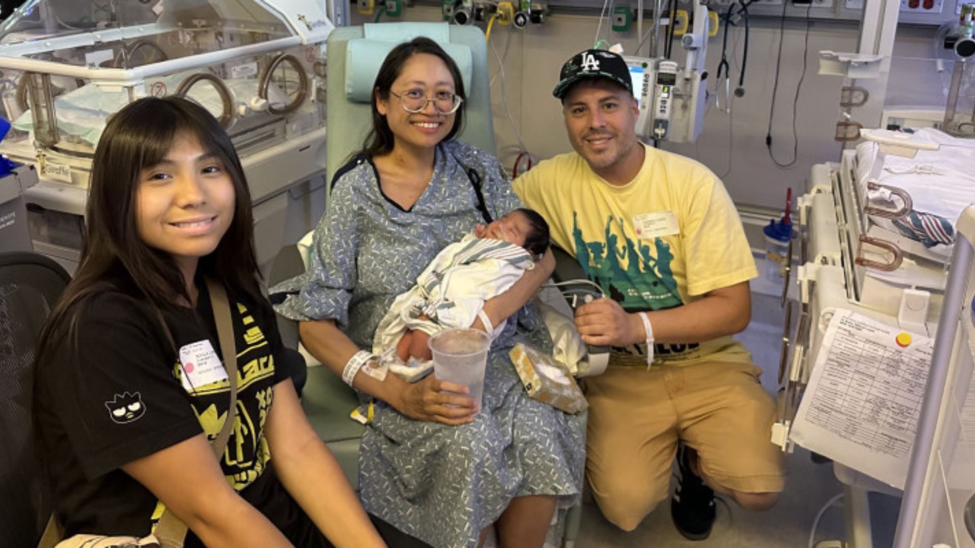 photo of three smiling individuals (a teen and two parents) in a hospital room. the mom, who is wearing a hospital gown, is at the center of the photo holding a baby wrapped in a blanket