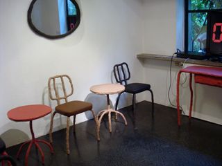 Left to right: An orange round top side table with five legs; a brown chair with four legs and a small back with a brown cushion; a beige table of the same style and a black chair of the same style.