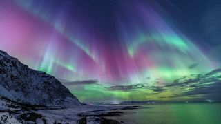 northern lights in Norway, appearing as vast ribbons or curtains of purple and green light, the entire sky is filled with color and is reflected in the water below.