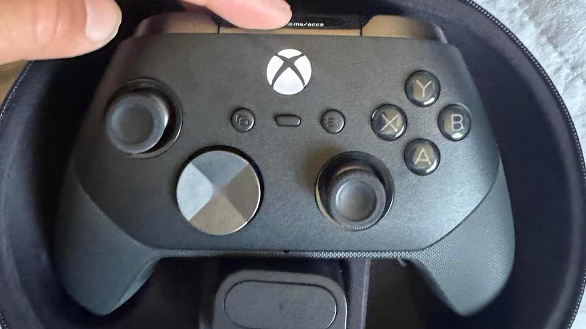 A black Xbox Elite wireless controller sits inside a round, zippered carrying case, featuring a metallic D-pad and standard gaming button layout.