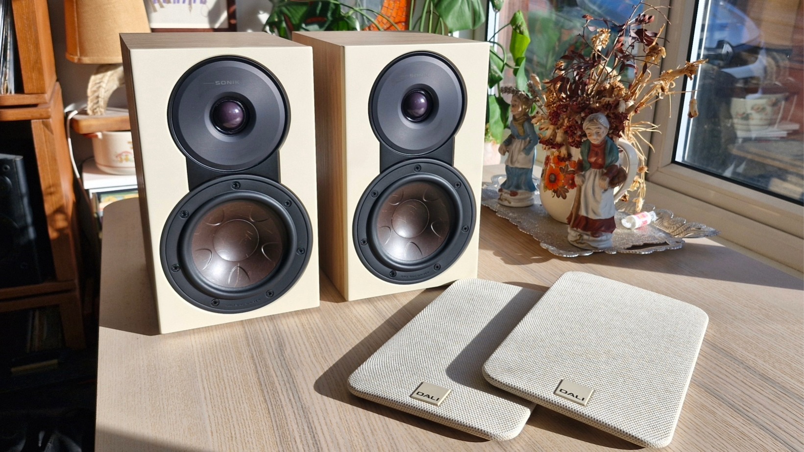 Dali Sonik 1 passive bookshelf speakers woofer and tweeter, with off-white grilles on a wooden surface in front.
