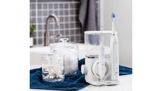 Image shows the Waterpik Complete Care 9.0 in a bathroom, alongside toiletries and a blue towel.