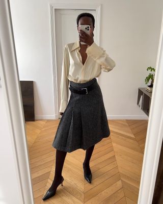 Sylvie wears an ivory silk blouse, gray pleated midi skirt, a black belt, black sheer tights, and black leather pumps.