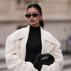 woman in white coat, black dress and sunglasses holding black leather handbag